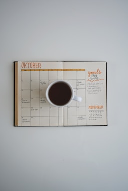 An overhead shot of an open planner displaying the months of October and November. A white coffee cup filled with black coffee is placed on top of the planner. The planner includes handwritten notes and goals for the month.