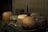 A collection of rustic clay pots, varying in size and shape, is arranged on a wooden surface. The pots exhibit an earthy color palette with shades of brown, green, and ochre, accompanied by signs of wear and age. A thatched or bamboo mat serves as a backdrop, adding to the rustic aesthetic.