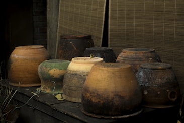 A collection of rustic clay pots, varying in size and shape, is arranged on a wooden surface. The pots exhibit an earthy color palette with shades of brown, green, and ochre, accompanied by signs of wear and age. A thatched or bamboo mat serves as a backdrop, adding to the rustic aesthetic.