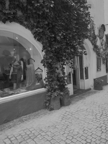 A charming storefront with a large arched window displaying mannequins wearing dresses. Lush ivy climbs up the white facade of the building. The cobblestone street adds a vintage feel. Barrels with plants decorate the entrance, enhancing the quaint atmosphere.