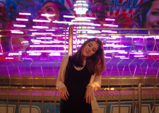 A woman stands in front of a bright and colorful amusement park ride with neon lights creating a dynamic and vibrant background. She has long hair and is wearing a black outfit with a light cardigan. Her expression is serene, with her head slightly tilted and eyes closed.