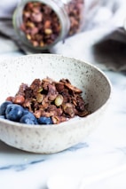 A textured ceramic bowl filled with crunchy granola mixed with nuts, accompanied by fresh blueberries. In the background, a jar filled with more granola is tipped over on a soft, grey cloth. The setting is on a smooth, white surface, possibly a countertop or table.