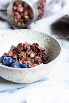 A textured ceramic bowl filled with crunchy granola mixed with nuts, accompanied by fresh blueberries. In the background, a jar filled with more granola is tipped over on a soft, grey cloth. The setting is on a smooth, white surface, possibly a countertop or table.