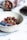 A textured ceramic bowl filled with crunchy granola mixed with nuts, accompanied by fresh blueberries. In the background, a jar filled with more granola is tipped over on a soft, grey cloth. The setting is on a smooth, white surface, possibly a countertop or table.