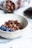 A textured ceramic bowl filled with crunchy granola mixed with nuts, accompanied by fresh blueberries. In the background, a jar filled with more granola is tipped over on a soft, grey cloth. The setting is on a smooth, white surface, possibly a countertop or table.