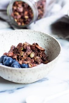 A textured ceramic bowl filled with crunchy granola mixed with nuts, accompanied by fresh blueberries. In the background, a jar filled with more granola is tipped over on a soft, grey cloth. The setting is on a smooth, white surface, possibly a countertop or table.