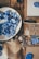 A collection of blue clay or wax seals and molds is scattered on a wooden table. The seals are in various sizes and shapes, including floral and geometric patterns. Brushes and crafting tools are also present, along with some metal or silver decorative pieces. Papers and other crafting materials are arranged around the table, hinting at an ongoing creative project.