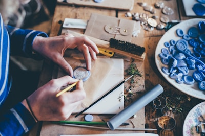 A person is engaged in crafting activities at a wooden table, working with blue clay or wax. They are holding a small round piece and a brush, surrounded by numerous carved or stamped clay items. The table is scattered with crafting tools, leaves, and materials, including envelopes, brushes, and a bowl of blue clay items.