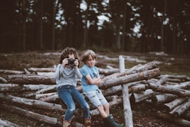 Two children are sitting outdoors on a pile of logs. One child is holding a camera, while the other is sitting with crossed arms. They are in a forested area with trees in the background, and the mood is relaxed and playful.