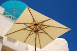 A large beige patio umbrella stands against a backdrop of white buildings, with vibrant blue skies and a turquoise umbrella visible in the background. The setting appears sunny and bright, suggesting an outdoor environment, possibly in a Mediterranean or coastal area.