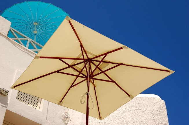 A large beige patio umbrella stands against a backdrop of white buildings, with vibrant blue skies and a turquoise umbrella visible in the background. The setting appears sunny and bright, suggesting an outdoor environment, possibly in a Mediterranean or coastal area.