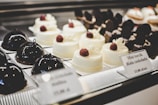 A selection of elegant pastries on display, including glossy dark chocolate dome-shaped cakes and smooth white chocolate desserts topped with raspberries. The desserts are arranged neatly on individual plates with labels describing each item.