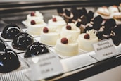 A selection of elegant pastries on display, including glossy dark chocolate dome-shaped cakes and smooth white chocolate desserts topped with raspberries. The desserts are arranged neatly on individual plates with labels describing each item.