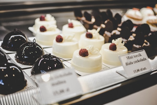 A selection of elegant pastries on display, including glossy dark chocolate dome-shaped cakes and smooth white chocolate desserts topped with raspberries. The desserts are arranged neatly on individual plates with labels describing each item.