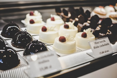 A selection of elegant pastries on display, including glossy dark chocolate dome-shaped cakes and smooth white chocolate desserts topped with raspberries. The desserts are arranged neatly on individual plates with labels describing each item.