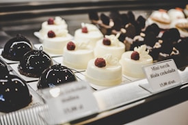 A selection of elegant pastries on display, including glossy dark chocolate dome-shaped cakes and smooth white chocolate desserts topped with raspberries. The desserts are arranged neatly on individual plates with labels describing each item.