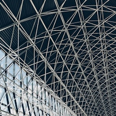A complex geometric pattern formed by intersecting steel beams creates an intricate lattice structure. The design is airy, with light filtering through the gaps, highlighting the architectural detail and symmetry.