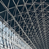 A complex geometric pattern formed by intersecting steel beams creates an intricate lattice structure. The design is airy, with light filtering through the gaps, highlighting the architectural detail and symmetry.