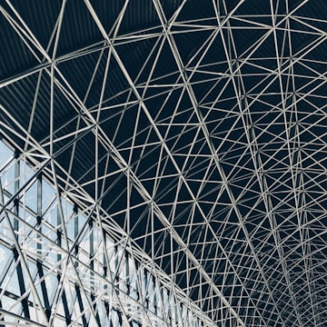 A complex geometric pattern formed by intersecting steel beams creates an intricate lattice structure. The design is airy, with light filtering through the gaps, highlighting the architectural detail and symmetry.