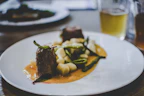 A gourmet dish is elegantly presented on a white plate. The meal consists of two pieces of tender meat garnished with fresh herbs, accompanied by white gnocchi and green vegetables, all served over a rich, creamy sauce. In the background, there's a blurred image of a drink, possibly a beer, in a glass.