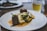 A gourmet dish is elegantly presented on a white plate. The meal consists of two pieces of tender meat garnished with fresh herbs, accompanied by white gnocchi and green vegetables, all served over a rich, creamy sauce. In the background, there's a blurred image of a drink, possibly a beer, in a glass.