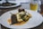A gourmet dish is elegantly presented on a white plate. The meal consists of two pieces of tender meat garnished with fresh herbs, accompanied by white gnocchi and green vegetables, all served over a rich, creamy sauce. In the background, there's a blurred image of a drink, possibly a beer, in a glass.