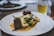 A gourmet dish is elegantly presented on a white plate. The meal consists of two pieces of tender meat garnished with fresh herbs, accompanied by white gnocchi and green vegetables, all served over a rich, creamy sauce. In the background, there's a blurred image of a drink, possibly a beer, in a glass.