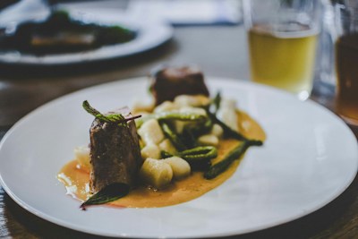 A gourmet dish is elegantly presented on a white plate. The meal consists of two pieces of tender meat garnished with fresh herbs, accompanied by white gnocchi and green vegetables, all served over a rich, creamy sauce. In the background, there's a blurred image of a drink, possibly a beer, in a glass.