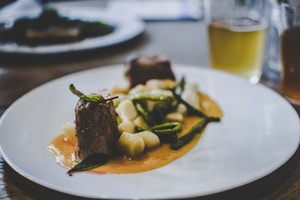 A gourmet dish is elegantly presented on a white plate. The meal consists of two pieces of tender meat garnished with fresh herbs, accompanied by white gnocchi and green vegetables, all served over a rich, creamy sauce. In the background, there's a blurred image of a drink, possibly a beer, in a glass.