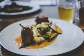 A gourmet dish is elegantly presented on a white plate. The meal consists of two pieces of tender meat garnished with fresh herbs, accompanied by white gnocchi and green vegetables, all served over a rich, creamy sauce. In the background, there's a blurred image of a drink, possibly a beer, in a glass.