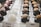 Rows of elegant pastries are displayed, with each one meticulously crafted. On the left, there are glossy, dark chocolate cakes topped with white cream and red decorations. In the center, domed white cakes are adorned with delicate, ivory-colored accents. To the right, dark glazed desserts hold macarons dusted with cocoa powder. The arrangement creates a sophisticated and appetizing presentation.