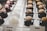 Rows of elegant pastries are displayed, with each one meticulously crafted. On the left, there are glossy, dark chocolate cakes topped with white cream and red decorations. In the center, domed white cakes are adorned with delicate, ivory-colored accents. To the right, dark glazed desserts hold macarons dusted with cocoa powder. The arrangement creates a sophisticated and appetizing presentation.