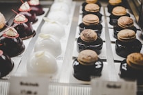 Rows of elegant pastries are displayed, with each one meticulously crafted. On the left, there are glossy, dark chocolate cakes topped with white cream and red decorations. In the center, domed white cakes are adorned with delicate, ivory-colored accents. To the right, dark glazed desserts hold macarons dusted with cocoa powder. The arrangement creates a sophisticated and appetizing presentation.
