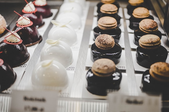 Rows of elegant pastries are displayed, with each one meticulously crafted. On the left, there are glossy, dark chocolate cakes topped with white cream and red decorations. In the center, domed white cakes are adorned with delicate, ivory-colored accents. To the right, dark glazed desserts hold macarons dusted with cocoa powder. The arrangement creates a sophisticated and appetizing presentation.