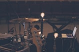 A dimly lit music scene features a drum set, a laptop, and some light bulbs on stands, creating a warm ambiance. The setting appears to be a rehearsal or recording studio with rows of empty seats in the background, suggesting a quiet, focused environment.