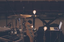 A dimly lit music scene features a drum set, a laptop, and some light bulbs on stands, creating a warm ambiance. The setting appears to be a rehearsal or recording studio with rows of empty seats in the background, suggesting a quiet, focused environment.