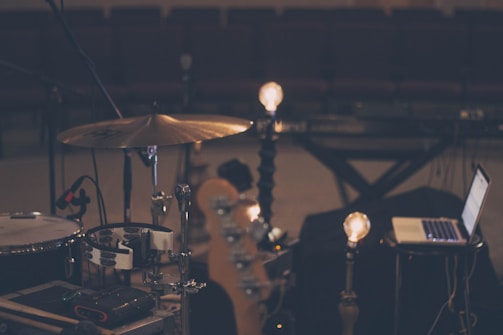 A dimly lit music scene features a drum set, a laptop, and some light bulbs on stands, creating a warm ambiance. The setting appears to be a rehearsal or recording studio with rows of empty seats in the background, suggesting a quiet, focused environment.
