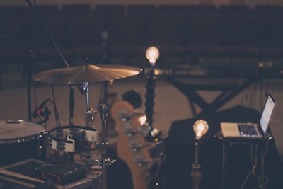 A dimly lit music scene features a drum set, a laptop, and some light bulbs on stands, creating a warm ambiance. The setting appears to be a rehearsal or recording studio with rows of empty seats in the background, suggesting a quiet, focused environment.