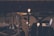 A dimly lit music scene features a drum set, a laptop, and some light bulbs on stands, creating a warm ambiance. The setting appears to be a rehearsal or recording studio with rows of empty seats in the background, suggesting a quiet, focused environment.