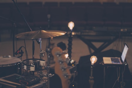 A dimly lit music scene features a drum set, a laptop, and some light bulbs on stands, creating a warm ambiance. The setting appears to be a rehearsal or recording studio with rows of empty seats in the background, suggesting a quiet, focused environment.