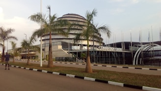 A modern architectural building featuring a large dome with multiple tiers. The foreground includes a row of palm trees lining a paved road with a person walking on it. Flags are visible to the side of the building.