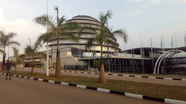 A modern architectural building featuring a large dome with multiple tiers. The foreground includes a row of palm trees lining a paved road with a person walking on it. Flags are visible to the side of the building.