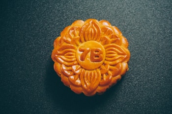 A round pastry with an intricate floral pattern embossed on its surface, set against a dark textured background. The pastry is a golden brown with a glossy finish, featuring the characters '7B' in the center.