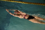 A swimmer is gliding through water with arms extended forward, wearing a black swimsuit with red straps and a swim cap. The reflection creates a shimmering effect on the water's surface, and a lane marker is visible at the top.