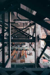 An industrial-style interior with exposed steel beams and concrete, featuring long wooden tables with benches hanging from the ceiling. The space has a rustic and modern contrast with visible pipes and ducts above.