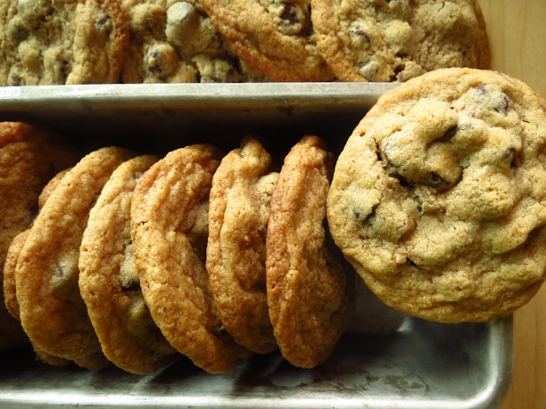 A batch of freshly baked chocolate chip cookies is neatly arranged in a metal tray. The cookies are golden brown with a slightly crispy texture around the edges and chunks of melted chocolate visible on the surface.