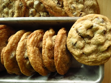 A batch of freshly baked chocolate chip cookies is neatly arranged in a metal tray. The cookies are golden brown with a slightly crispy texture around the edges and chunks of melted chocolate visible on the surface.