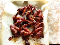 A pile of glazed chicken wings arranged on a sheet of parchment paper, with a slightly charred, sticky texture and a glossy red-brown color, suggesting they have been cooked with a rich barbecue sauce.