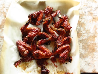 A pile of glazed chicken wings arranged on a sheet of parchment paper, with a slightly charred, sticky texture and a glossy red-brown color, suggesting they have been cooked with a rich barbecue sauce.