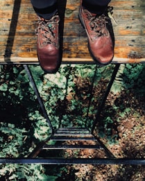 A pair of rugged brown leather boots stand on a wooden platform. Below is a metal ladder leading down to a forest floor covered with green foliage and dappled sunlight.